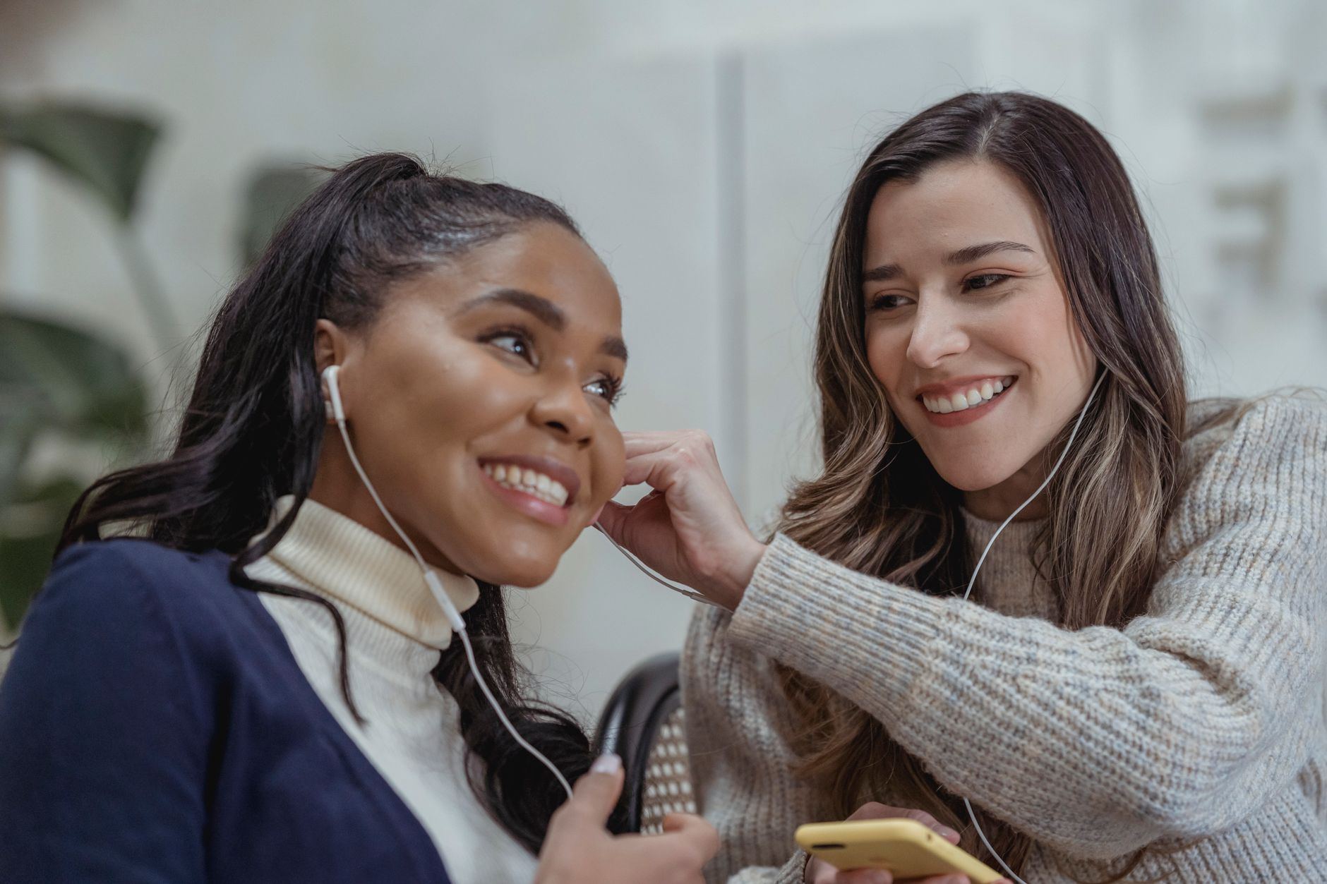 Cheerful multiethnic women sharing music in earphones and smiling on blurred background of apartment